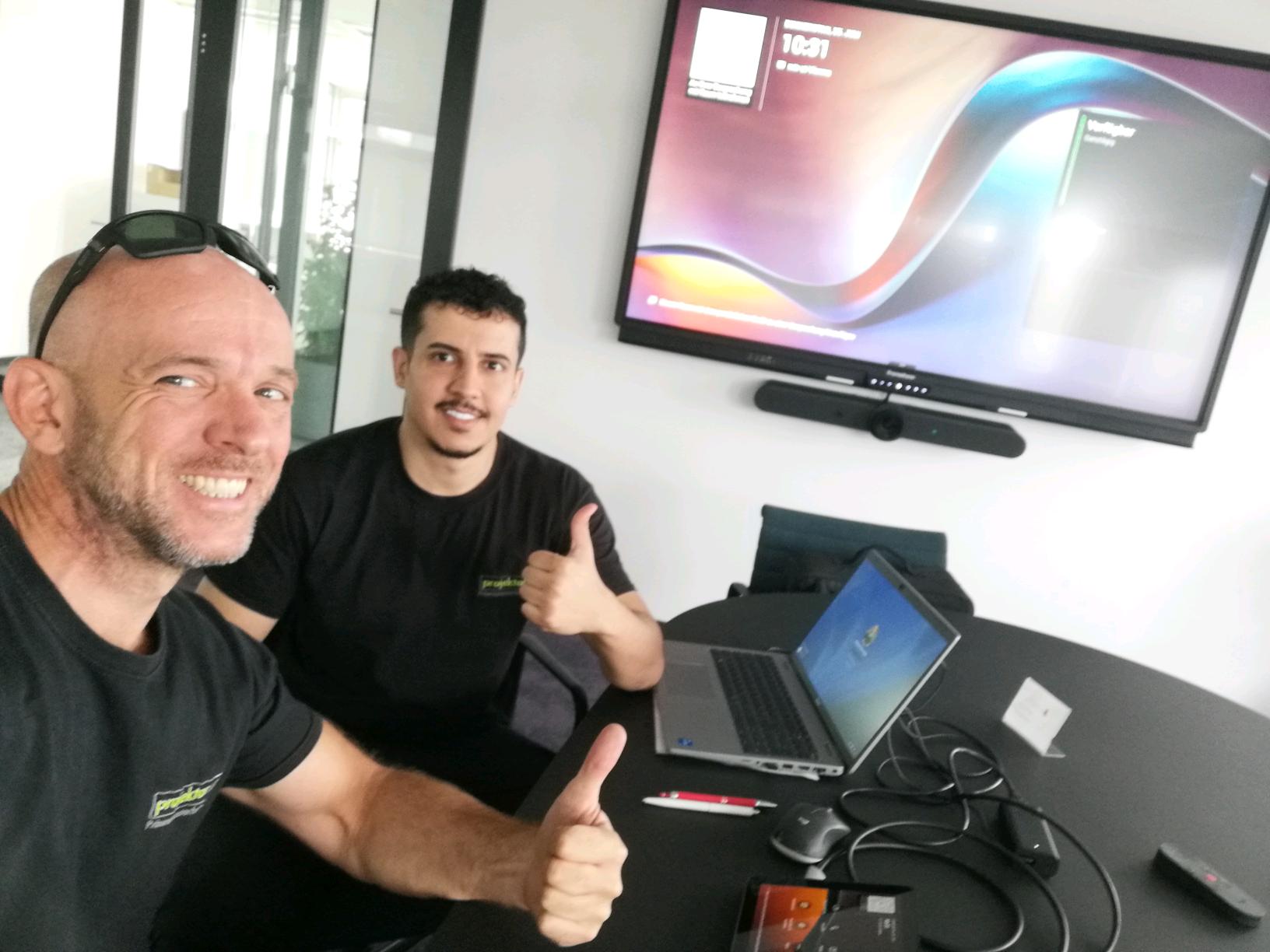 Zwei Techniker von projektor.at installieren und testen ein interaktives Display in einem modernen Konferenzraum. Beide tragen projektor.at-Shirts und zeigen Daumen hoch, während ein Laptop und Verbindungskabel auf dem Tisch liegen.